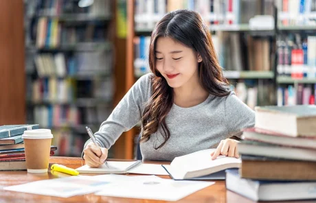 A woman is sitting at a library, taking notes from a book.