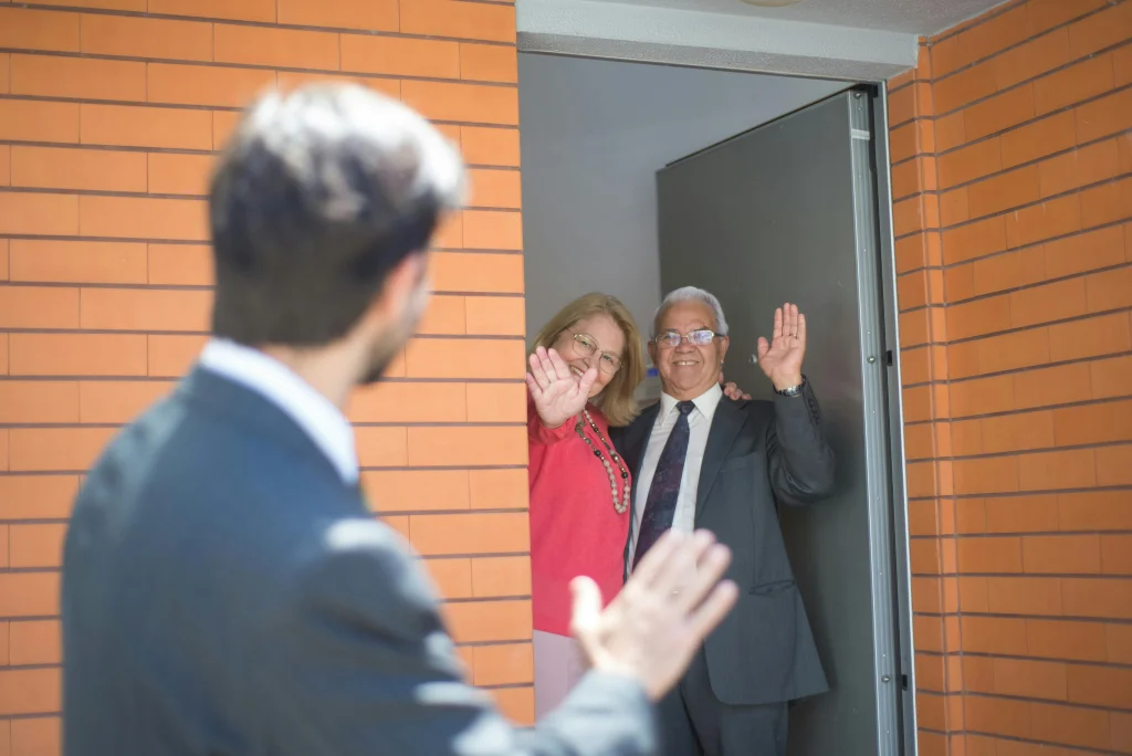 A man and at a woman wave at a man from their doorway.