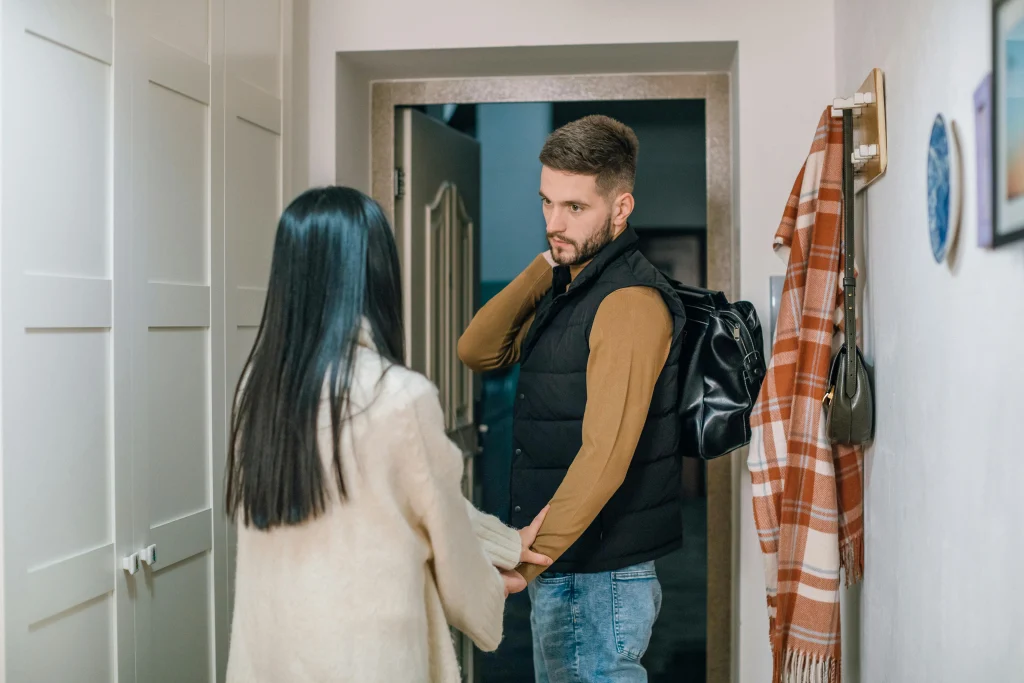 A woman holds a man’s hand while he carries a bag and stands before an open door.