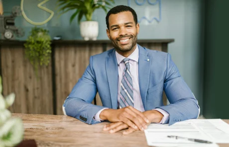 A man in a suit smiles with his hands crossed over his desk.
